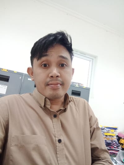 An adult male is depicted in a frontal, head-and-shoulders portrait, located within an indoor setting identified as Semarang, Indonesia. The individual has dark, styled hair and minimal facial stubble, gazing directly into the camera with wide eyes and slightly parted lips. He is wearing a light brown, long-sleeved button-up shirt with dark buttons and a visible left chest pocket. The background features plain white walls. To the subject's left, two grey metal filing cabinets are partially visible; the leftmost cabinet displays an illegible label. To the subject's right, a white-framed window is present, and below it, a stack of folded, multi-colored fabric items, possibly textiles or clothing, is visible, topped with several papers.