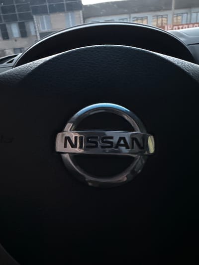 This is a close-up, angled shot of a Nissan emblem on a car's steering wheel. The emblem, with its silver circular border and the word "NISSAN" prominently displayed vertically in the center, is the main subject. The steering wheel appears to be made of dark, textured material, likely leather. The background is blurred and shows a view through a window, revealing a cityscape with buildings. The buildings have a pattern of windows and appear to be urban structures. The lighting suggests it could be daytime, given the visibility of the outside scene. The image is captured from within the car, looking towards the steering wheel and the view outside. There is no activity or event taking place, as the image is a static shot of an object and its surroundings. The visible text is "NISSAN". The location context of Shatava, Ukraine, is provided but not visually evident in the image itself.