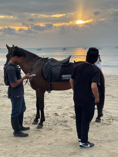 An overcast sunset casts a warm glow over Jimbaran Beach in Kuta Selatan, Indonesia. Two men stand on the sandy shore next to a brown horse. The man on the left, wearing a dark t-shirt, blue jeans, and tall black boots, is looking at his phone. The man on the right, dressed in black pants, a black t-shirt, and a black baseball cap, stands with his back to the camera. The horse is saddled with a black saddle, and a saddle pad with the text "JIMBARAN HORSE STABLE" is visible. In the distance, the ocean is dotted with traditional Balinese fishing boats, and the sun's rays break through the clouds, reflecting on the water. The sand is light brown and textured with footprints.