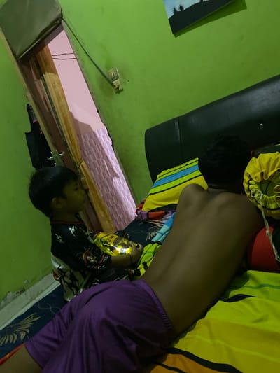 In the city of Ta', Indonesia, an indoor scene depicts two individuals in a dimly lit room. The room features green walls and is furnished with a bed. On the bed, a person is lying on their back, facing away from the camera, with their torso visible. They are wearing purple shorts. Next to them, a child stands and holds a yellow object, possibly a toy or snack packaging. The background reveals a doorway with a pink curtain and a patterned bedspread. The overall impression is a casual, domestic setting.