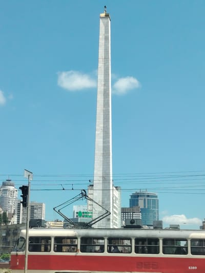 This image captures a street scene in Kyiv, Ukraine, under a bright, clear blue sky with a few wispy clouds. The dominant feature is a tall, slender, white obelisk, likely a monument, topped with a golden pinnacle. In the foreground, a red and cream-colored tram is moving, its pantograph extended to draw power from overhead wires. The tram has multiple windows, and through them, several passengers can be seen.
Behind the tram and to the left of the obelisk, a cluster of modern buildings rises, including a distinctive round-topped skyscraper with a glass facade, and other high-rise structures. A traffic light is visible on a pole, and more overhead wires crisscross the frame, indicating an urban environment with active public transportation. Some faint text is visible on the side of the tram, and on some of the buildings in the background. The overall impression is a sunny day in a bustling city, showcasing both historical landmarks and modern architecture.