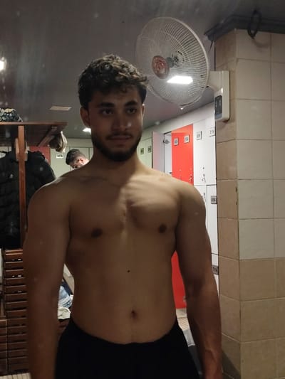 A shirtless man with curly hair and a beard stands in a locker room, posing in front of a mirror. He is wearing black athletic shorts. In the background, there are lockers, some of which are red and white. A fan is mounted on the wall above the lockers. Another person is partially visible in the reflection of the mirror, in the background of the image.