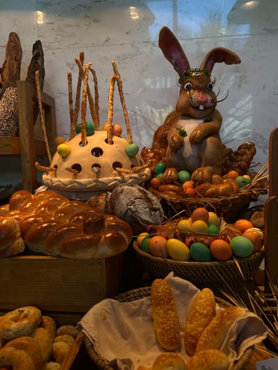 The image depicts a decorative display of Easter-themed items, likely set up in a commercial establishment in Nusa Dua, Indonesia.
Foreground:
A large, cartoonish rabbit figure with a floral wreath around its head is prominently displayed. A decorative object with holes filled with colored eggs and bread sticks. Several loaves of braided bread and various other breads are arranged.
Background:
Baskets filled with colorful Easter eggs and various bread types. The overall arrangement is intended to be festive and celebratory, targeting customers of the establishment.
Setting:
The image was likely taken indoors, indicated by the lighting and the marble-like tile floor. The scene suggests a bakery or a restaurant setting, and the decoration clearly targets Easter festivities.