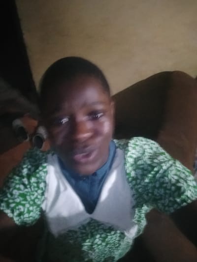 A person is looking at the camera, with their face filling the frame. They are wearing a green and white floral patterned shirt with a blue collar visible underneath. The background is dark and indistinct, suggesting an indoor setting. The image is blurry and low resolution. The location within Shangisha, Nigeria cannot be determined from the provided image.