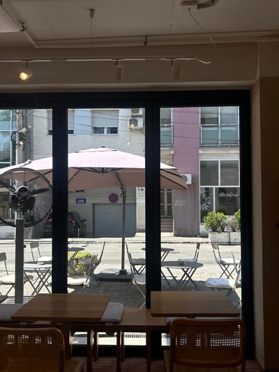 The image shows an interior view looking out through a large window onto a street scene. The perspective is from inside a cafe or restaurant.

The scene outside depicts a street with several tables and chairs set up, suggesting an outdoor seating area. Umbrellas are deployed, providing shade. Buildings line the street, with some featuring balconies and windows. One building has a sign that reads "Hairlove." There are cars visible on the street, and a fan is positioned inside the cafe, near the window. The time of day appears to be daytime, with bright sunlight casting shadows.

Visible text includes "Hairlove" on a building sign, and some graffiti on a wall. There are also signs on the street, including a red circular sign with a diagonal line and a blue sign with a symbol, possibly indicating parking regulations.

The overall impression is of a European street scene, likely in a city, with a relaxed atmosphere during the day. The indoor elements, such as the tables, chairs, and fan, suggest a functional commercial space. The lighting indicates it's a sunny day.