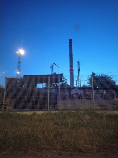 The image shows an industrial area at dusk in Pilsen, Czechia. A tall, cylindrical chimney with red lights near its top is prominently featured in the center-right of the frame. To the left of the chimney, a lattice tower with bright floodlights illuminates the surrounding area. In the foreground, a chain-link fence encloses the industrial site, behind which are some covered structures and what appear to be stacked materials wrapped in plastic. Trees are visible behind the fence and around the industrial structures. The sky is a deep blue, indicating twilight. The ground in front of the fence is covered in dry grass. There are no people visible in the image.