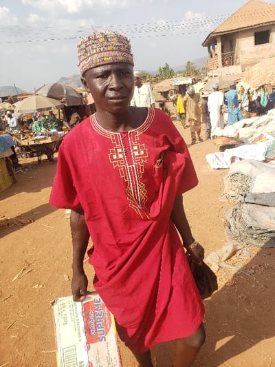 An outdoor scene in a market area of Adonkolo, Nigeria, depicts a male individual in the foreground and market activity in the background. The central figure is a dark-skinned male, appearing middle-aged, standing on a reddish-brown dirt path and facing directly towards the camera. He wears a red short-sleeved tunic adorned with white and gold geometric embroidery on the chest, and a multi-colored, patterned skullcap. His skin appears glistening. He holds a white and red carton labeled "SUPREME" and "40 x 120g" from underneath with his left hand. In his right hand, he carries a dark-colored bag or pouch; a gold-colored bracelet is visible on his right wrist.
In the left background, several market stalls with light-colored umbrellas providing shade are visible, displaying various goods. Multiple individuals are present among these stalls. Overhead, power lines span across the upper left sky. In the right background, a two-story building with a reddish-brown roof and exposed construction appears. Further along the dirt path, more people are observed, and piles of bundled materials are on the ground near the frame's right edge. Distant hills or mountains are visible on the horizon under a partly cloudy sky. The entire ground surface is reddish-brown dirt.