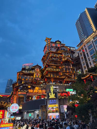 The image shows Hongyadong, a popular tourist attraction in the Yuzhong District of Chongqing, China. The architecture features traditional Chinese elements with multi-tiered wooden structures adorned with red lanterns and bright neon signs in Chinese characters. The buildings are lit up, suggesting it is dusk or night. Crowds of people are visible on the street level, indicating the location's popularity. Modern skyscrapers are visible in the background, contrasting with the traditional buildings.