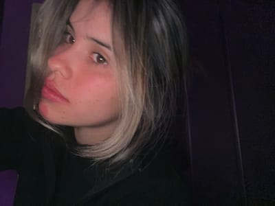 The image is a close-up, indoor, self-portrait of a young woman with fair skin, freckles, and blonde and brown hair. She is wearing a black top and is positioned in a dark room with purple walls. Her face is turned to the side, and her eyes are looking upwards and to the left. The lighting is dim, casting shadows on her face and the background. The woman appears to be in her late teens or early twenties. There are no other individuals or objects clearly visible in the frame. The scene does not provide any information that would allow for the identification of a specific location within Sumalao, Argentina.