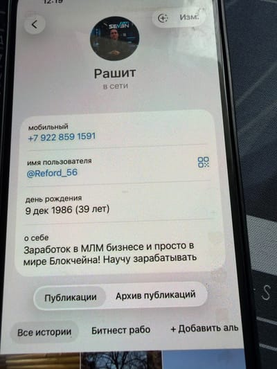 The image is a close-up of a smartphone screen displaying a profile page. The profile belongs to a user named "Рашит" (Rashit), who is currently "в сети" (online). The profile shows a circular profile picture with a person wearing a suit and a logo that appears to say "SEVAN". Below the name, the mobile number is listed as "+7 922 859 1591". The username is "@Reford_56". The date of birth is "9 дек 1986" (December 9, 1986), and the age is indicated as "(39 лет)" (39 years). The "About me" section states "Заработок в MLM бизнесе и просто в мире Блокчейна! Научу зарабатывать" (Earning in MLM business and simply in the world of Blockchain! I will teach you to earn). There are buttons for "Публикации" (Publications) and "Архив публикаций" (Publication archive). Further down, there are options for "Все истории" (All stories), "Битнест рабо" (Bitnest work), and "+ Добавить аль" (+ Add other). A QR code icon is also visible. The background of the phone is dark and textured. The image is taken from a slightly angled perspective, suggesting it is being held in hand. There is no indication of the city of Orenburg, Russia, in the provided image.