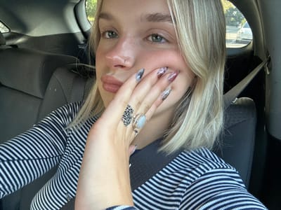A young woman is depicted in the front passenger seat of a vehicle, situated in a city in Victoria, Canada. She has shoulder-length blonde hair, light-colored eyes, and a small hoop nose ring on her left nostril. Her lips are slightly parted, enhanced with a light pink or nude cosmetic. She wears a long-sleeved shirt with horizontal dark blue and white stripes, secured by a dark seatbelt across her chest. Her right hand is positioned near her chin and mouth, displaying long, manicured fingernails with an intricate marbled design in shades of grey, white, and reddish-pink. Two rings are visible on her right hand: an ornate silver-toned ring with a complex design on her ring finger, and a silver-toned ring with a white cabochon stone on her middle finger. The vehicle's interior features dark grey upholstery. Through the passenger window, green foliage and the side of a light-colored vehicle are visible in daylight.
