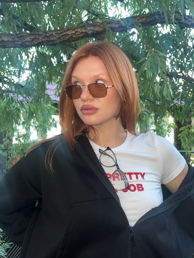 A young woman with auburn hair and sunglasses is pictured outdoors in Chaykovsky, Russia, with a leafy tree in the background. She is wearing a white t-shirt with red lettering that reads "PRETTY JOB" and a black zip-up jacket. A pair of glasses is draped over her t-shirt. The image is framed from the chest up, focusing on the woman's face and upper body.