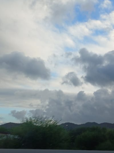 The image depicts a landscape scene under a cloudy sky. In the foreground, a dark, blurry green bush or tree line stretches across the lower portion of the frame. Behind this foliage, a dark, undulating mountain range is visible. A single, small, bright light, possibly a street lamp, is illuminated on the mountainside. The sky is filled with a mix of light and dark gray clouds, with patches of blue sky showing through. The overall impression is a natural environment, possibly a rural or mountainous area on the outskirts of a city. Based on the description of Porlamar, Venezuela, this scene could represent the outskirts of the city, where the natural landscape meets the urban environment.