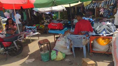 This image captures an outdoor market scene in Moshi, Tanzania, under bright daylight. In the left foreground, a woman with dark hair, dressed in a floral top and a plaid skirt, is seated sideways on a red motorcycle, looking down. Behind her, a man in a white t-shirt stands partially visible near a display. To the immediate right of the motorcycle, a table holds multiple stacks of folded garments. A red market umbrella provides shade over this section.
In the center, a small red table displays a red garment and other clothing items. On the ground nearby, a green bucket contains a wooden implement, and a small cushioned stool is present. Several large sacks, including white and green ones, are situated on the dirt ground.
On the right, a woman wearing a red shirt and a patterned skirt is seated on a light-blue plastic chair, facing away from the viewer. She is actively sorting through clothing items arranged on an orange table in front of her. A large white sack with a yellow liner is positioned beneath this table. A green market umbrella provides overhead shade on the right.
The background features numerous market stalls with various clothing items piled on tables and hanging from makeshift structures. The ground throughout the scene is composed of packed dirt or gravel.