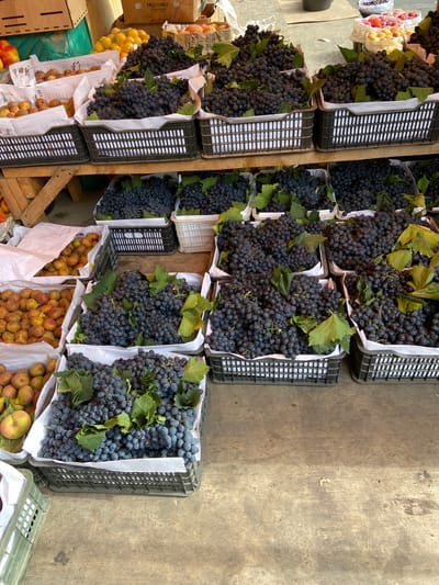 An outdoor fruit stall in Dushanbe, Tajikistan displays an abundance of fresh produce. Several crates are filled with dark purple grapes, some still adorned with green leaves. Interspersed among the grapes are other fruits, including what appear to be plums or apricots in various stages of ripeness, and a single pale green and yellow apple. In the background, stacks of cardboard boxes, some with partial text legible as "FRUTADELI," suggest commercial packaging. The stall is set up on a wooden surface, with a slightly worn, textured appearance. The overall impression is one of a vibrant, local market scene.
