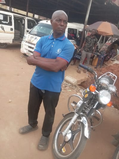 This image captures a street scene in Nnobi, Nigeria. In the foreground, a bald, dark-skinned male, approximately 40-50 years old, stands centrally with his arms crossed, wearing a blue polo shirt with a "KYMOTO C" logo, black pants, and dark grey clogs. He faces slightly right of the camera with a neutral expression. To his right, a silver and black motorcycle with a round headlight and orange turn signals is parked. The ground is an unpaved, reddish-brown dirt surface.
In the mid-ground, a white passenger van with its side door open reveals stacks of tires inside on the left. Another white van is visible behind the main subject, with a person's face partially visible through its window. Further right, a dark-skinned male in a patterned short-sleeved shirt and light pants stands near a brown market umbrella. Several individuals are partially visible behind the vans and umbrella, some seated on plastic chairs. A yellow container and various debris are scattered on the ground. Overhead, a dark structure suggests a covered market area. A partially obscured sign near the umbrella reads "POS & TRANSFER."