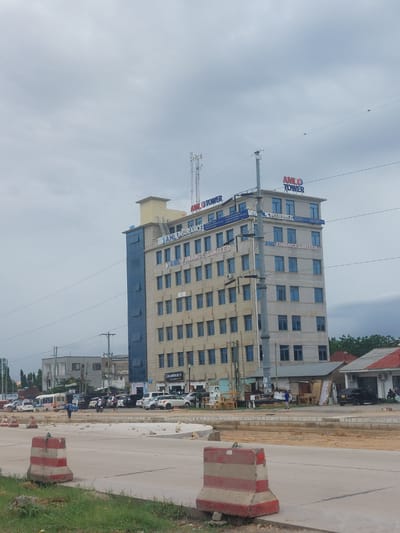 The image depicts a multi-story building, identified as "AMIL Tower," in Dar es-Salaam, Tanzania. The building has a combination of grey and blue facade elements. Street-level traffic and infrastructure are visible in front of the building. A section of roadway with painted barricades is also present. The sky is overcast.