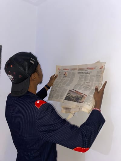 An individual is depicted reading a newspaper, seen in a rotated orientation with their head facing left. The person, who appears to be male, is wearing a black mesh baseball cap and a dark pinstriped jacket adorned with distinctive red, circular elbow patches. A black wristband is visible on their left wrist, and a pink wristband with the text "FOR JESUS" is on their right arm. They hold an open newspaper with both hands, and their right index finger is pointing at a section of the page. The background is a plain, light-colored wall or surface, indicating an indoor setting.
Visible text on the newspaper includes headlines such as "Let's talk about premenstrual disorders", "Responding to reality", and "Emerging roles, agencies of Education ministry". There is also a large "H" and "0000" visible. The person's expression is not fully clear due to the angle, but they appear focused on reading. The scene takes place in Joma, Ghana.