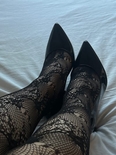 The image shows a pair of legs clad in patterned black tights and black patent leather high heels resting on a white surface. The feet are crossed at the ankles. The background is a crumpled white cloth, possibly a bed sheet. The location is not discernible from the image and cannot be identified as Novi Sad, Serbia or any other location.