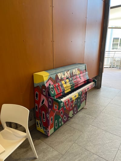 This media file captures a vibrant, custom-painted public piano situated in an indoor or covered public space in Kelvin Grove, Australia. The piano is adorned with a colorful, whimsical mural depicting numerous stylized houses or buildings in various shades of red, green, pink, and yellow, creating a cityscape design. Visible text on the side of the piano includes "PLAY ME" and "PIANO YOURS," indicating its interactive nature.
To the left of the piano is a large, unadorned reddish-brown wall or paneling. The floor beneath and to the right of the piano is covered with light-colored, square tiles. In the bottom right corner, a portion of a white, modern plastic chair is visible, suggesting a seating area. The background, particularly in the upper left, shows elements of a building structure, including windows and railings, under bright, natural daylight. The setting appears to be a clean and well-maintained public area, possibly an atrium or wide corridor. There are no people visible in the image.