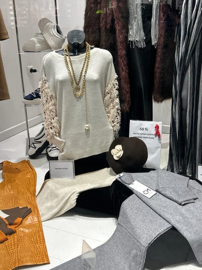 The image depicts a retail display, likely a shop window, showcasing various clothing items and accessories. The scene features multiple mannequins and arranged garments, suggesting a fashion store in Koridallos, Greece, as indicated by visible Greek text.
In the foreground, a cream-colored long-sleeved top or sweater with textured, possibly fringed, shoulder embellishments is displayed on a mannequin bust. A gold layered necklace with a circular pendant and a small pearl adorns the top. To its left, a pair of white sneakers are visible on a lower display surface. To the right, a dark brown cloche hat with a white floral appliqué is featured.
Further into the display, a grey sweater or jacket is draped, with a white tag bearing the "CM" logo attached. Adjacent to this, dark brown fur-like material is arranged horizontally, possibly part of a coat or trim, along with reflective silver fabric strips. The display surfaces are glossy and reflective, showing slight distortions of the items.
Several informational signs are present. A white sign with a red ribbon prominently displays "-50%," indicating a discount. Below this, Greek text reads "ΠΑΛΤΟ-ΓΟΥΝΕΣ-ΖΑΚΕΤΕΣ" (Coats-Furs-Jackets) and "ΕΙΔΟΝΤΑΙ ΠΑΡΑΓΓΕΛΙΕΣ" (Orders are accepted/seen). Another smaller sign clearly shows "ΜΠΟΤΕΣ 45.00€" (Boots 45.00€). The display is static, designed to showcase merchandise for sale. No people are visible, and the time of day is not discernible from the indoor lighting.