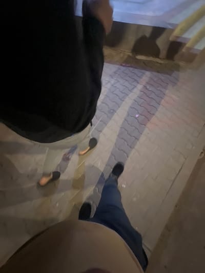 The image, taken at night, depicts a street scene with three individuals. The location is suggested to be in a city, as indicated by the tiled sidewalk. Two individuals are partially visible; one is wearing dark clothing, and the other is wearing blue jeans and light-colored clothing, possibly tan or beige. The third individual's shoes are visible near the center of the image. The scene is dimly lit, and the shadows cast by the individuals are visible on the sidewalk.