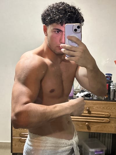 A shirtless man with curly hair and a towel wrapped around his waist takes a selfie. He holds a lilac iPhone in his left hand, partially obscuring his face. He poses with a flexed right arm, showcasing his muscles. The background features a wooden cabinet, various toiletries, and a plastic storage container. The image has been rotated and appears to be lit by a soft, natural light.