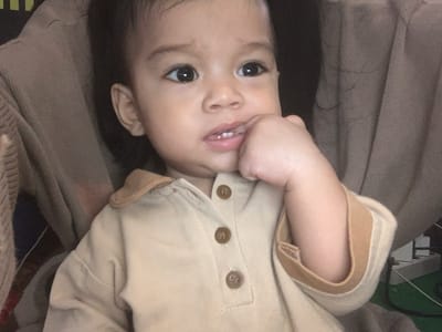 A close-up, head-and-shoulders shot focuses on a young child, appearing to be less than two years old, sitting and possibly being held. The child has dark hair and large, dark eyes. One hand is raised to the child's mouth, with fingers touching the lips and visible teeth. The child is wearing a light brown collared shirt with three visible brown buttons down the front. The background is soft and out of focus, showing textures of fabric, possibly a sweater or blanket, in neutral tones of brown and beige. The overall lighting is dim. The location in Sungai Meriam, Indonesia, is not discernible from the visual information in the image.