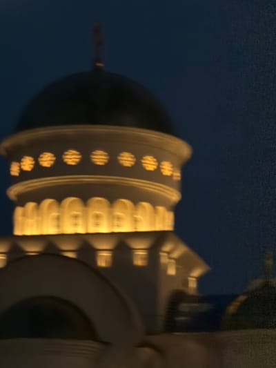 The image shows a close-up, blurred view of a church dome and facade at night. The dome is dark with a golden cross on top. The facade is illuminated by warm yellow lights, highlighting decorative architectural features. The sky is a deep, dark blue. The overall impression is of a religious building in an urban setting at night, with a focus on its illuminated architecture. The location is identified as the Church of St. John Vladimir in Bar, Montenegro.