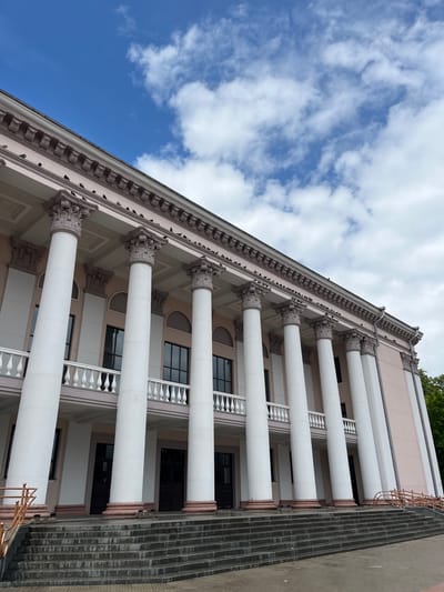 The image shows a wide shot of the building housing the Hrodna Regional Drama Theatre, located in Hrodna, Belarus. The building features a classical facade with a colonnade of white Ionic columns supporting a light pink entablature. A white balustrade runs along the second-story balcony. The sky above is a bright blue with scattered white clouds. Several pigeons are perched on the decorative molding along the top of the building. In the foreground, a wide set of stone stairs leads up to the entrance. The overall impression is one of stately architecture under a clear sky.