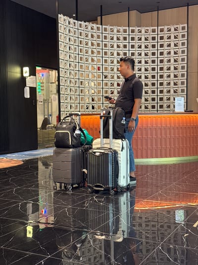 The image shows a man with short, dark hair leaning over a luggage cart, looking at his phone. He is wearing a dark t-shirt and blue jeans. The luggage cart holds several suitcases, including a large black one, a white one, and a smaller black and silver one. There is also a black backpack and a green bag on the cart.
The scene appears to be indoors, possibly a hotel lobby or an airport, given the presence of luggage and a sign that says "RECEPTION COUNTER." The background features a textured white wall with square openings, a wall with horizontal orange slats, and a dark, reflective floor with a marble-like pattern. The lighting suggests it could be daytime.
In the distance, a black door is visible, and above it, a panel with various lights and buttons, potentially a control panel or a reception area. The overall atmosphere seems to be one of transit or waiting. The man's posture suggests he might be checking information or communicating. The image is taken with a vertical orientation.