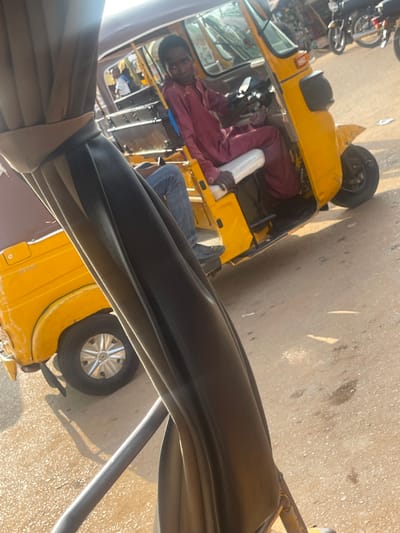 In Galadimawa Village, Nigeria, a young boy, dressed in a maroon kaftan and blue jeans, sits at the helm of a yellow auto-rickshaw, likely engaged in its operation or in transit. The vehicle, a common mode of transport in the region, is partially obscured by the foreground, which appears to be the interior of another vehicle or structure, possibly a canopy or a seat, covered in dark fabric with a beige trim. The scene unfolds on a dusty, sun-drenched street, with the bright sunlight casting distinct shadows. In the background, other auto-rickshaws and motorcycles suggest a busy urban or semi-urban environment. The boy's gaze is directed forward, hinting at his focus on the road ahead.