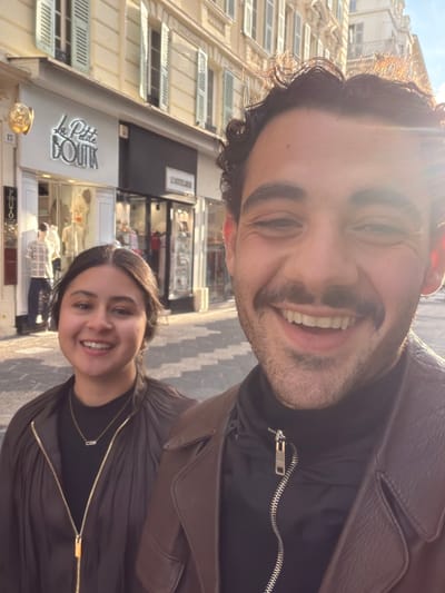 A selfie-style photograph captures two smiling individuals on a city street in Nice, France, specifically 5 Rue Massena. In the foreground on the right, a male with dark curly hair, a mustache, and stubble wears a dark brown jacket over a black zipped top, smiling broadly at the camera. To his left and slightly behind, a female with dark hair and dark eyes wears a dark, possibly pleated or ruched, zipped top and a gold chain with an elongated pendant, also smiling directly at the camera. The background features light-colored, multi-story buildings with louvered shutters on the windows. A shop named "La Petite BOUTIK" with illuminated lettering is visible behind the female, showcasing mannequins in its window. Strong sunlight from the upper right creates a lens flare and highlights the male's hair, casting shadows consistent with a bright outdoor setting.