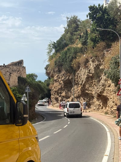 This media file depicts a street scene in Benalmádena, Spain, likely during the daytime under clear skies. The main subjects are several people and white vans on a winding road.

In the foreground, the bright yellow hood and side mirror of a vehicle are visible, suggesting the perspective is from inside or alongside another car. The road curves to the right, bordered by a sandy-colored rock face on the left and a paved area with a red brick border on the right.

Several people are standing near the white vans. One van, closer to the foreground, has its rear door open, and a few individuals are gathered around it. Further up the road, another white van is parked, and more people are visible standing near it and on the sidewalk. One person is seated on a low wall.

The rock face is covered in patches of green vegetation. A streetlamp extends from the rock face over the road. Traffic signs are visible on poles, including a red and white circular sign and a rectangular sign with the number "2".

The overall scene suggests a casual gathering or a pause in travel. The bright sunlight indicates it is daytime, and the clear sky suggests fair weather. The location is identifiable as Benalmádena, Spain, based on the provided context.