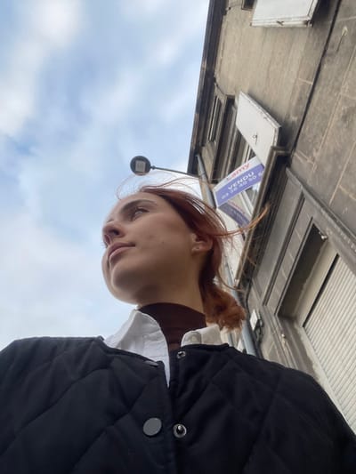 A low-angle shot captures a young individual, likely female, with reddish-brown hair pulled into a low ponytail, looking upwards and to the left. She wears a dark brown turtleneck beneath a white collared shirt, topped with a black quilted jacket featuring visible dark snaps. A silver stud earring is present on her left ear. The upper left portion of the frame shows a partly cloudy sky. To the right, a multi-story building with a light gray, textured facade dominates the background. A black streetlight fixture extends from the building. Affixed to the building, below an upper window with closed shutters, is a blue and white real estate sign. The sign, partially obscured, displays "Lamy," "VENDU" (French for "SOLD") in large red letters, and a phone number starting with "03 26 40 50 6". A lower window features a partially open, off-white shutter. The phone number prefix "03 26" on the sign identifies the location as Reims, France.