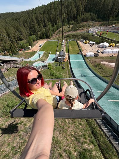 The image depicts a woman with short, red hair and sunglasses taking a selfie on a ski lift with a child wearing a tan hat and yellow shirt. They are seated on a black chair lift with metal safety bars. The background reveals a ski jump facility. The green run of the ski jump is visible below, along with trees and buildings. The location is likely Прикамский, Russia, based on the ski jump facility.