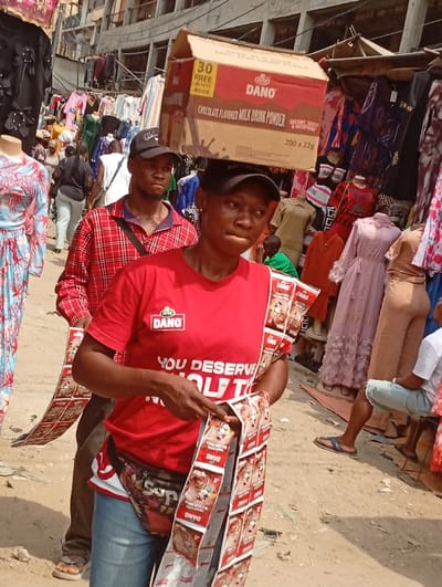 The image depicts a bustling outdoor market scene in Fegge, Nigeria, under bright daylight. In the immediate foreground, a person wears a red DANO-branded t-shirt with text "YOU DESERVE..." visible, blue jeans, a black baseball cap with a white logo, and a "SUP"-branded fanny pack. This individual is balancing a large, rectangular brown cardboard box labeled "DANO Chocolate Flavored Milk Drink Powder," prominently displaying "30 FREE SACHETS INSIDE," on their head. They are holding and examining multiple strips of DANO sachets with both hands.
Directly behind this individual, a male subject wearing a red and black plaid long-sleeved shirt and a dark baseball cap with a white logo walks forward. A dark strap crosses his left shoulder, and a strip of DANO sachets is draped over his left forearm.
The background is densely populated with market stalls and activity. Numerous items of clothing, including various dresses and garments in colors such as blue, pink, beige, red, and black, are displayed on mannequins and hanging from racks and structures. A multi-story concrete building with visible balconies and clothes hanging from lines is present in the upper background, beyond the immediate market stalls. Multiple other individuals are visible throughout the background, walking or browsing merchandise. The ground surface appears unpaved, dusty, and uneven.