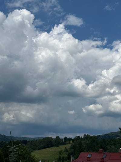 The image depicts a cloudy sky over a landscape featuring rolling hills covered in dense green trees. In the foreground, the roof of a red building with dormer windows is visible. The scene is a view from a higher elevation, looking down onto the treed slopes and towards a building. The sky is dynamic, with large, puffy white clouds in the upper portion gradually transitioning to darker, more ominous gray clouds lower down, suggesting an approaching storm. The overall impression is one of a rural or semi-rural setting in Wisła, Poland, with a focus on the interplay between the dramatic sky and the verdant landscape. There are no people or active events shown.
