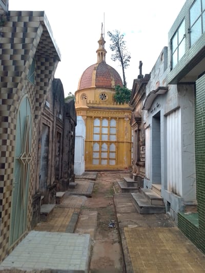 The image shows a view down an alley in the Recoleta Cemetery in Asuncion, Paraguay. The alley is lined with mausoleums on both sides. The mausoleums are made of various materials, including tile, stone, and concrete. Some of the mausoleums have windows and doors. In the center of the image, there is a large mausoleum with a dome. The ground is paved with stone and tile. There is no visible activity.