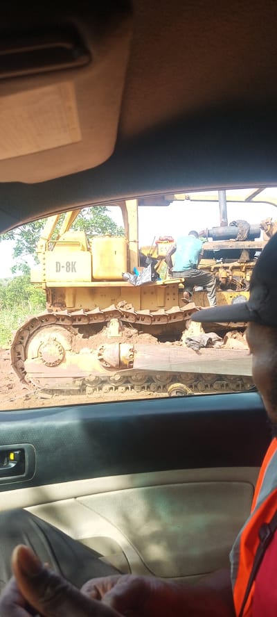 The image captures an outdoor scene from the interior of a car, looking out the passenger window. In the immediate foreground, part of a car's dashboard, a door panel with a handle, and the arm and shoulder of a person wearing an orange safety vest and a black cap are visible.

The central subject is a large, yellow heavy-duty bulldozer, identified by the text "D-8K" prominently displayed on its side. Its track assembly is heavily covered in mud and dirt, suggesting recent use in challenging terrain. Two men are present on or near the bulldozer. One man, wearing a light blue t-shirt and dark trousers, is sitting on a part of the machinery, facing away from the viewer, appearing to be engaged in an inspection or repair task. Another individual is partially visible further back on the bulldozer. Various items, including a silver-colored bag and an object with "CA" visible, are placed on the bulldozer's platform.

The background features green trees and foliage, indicating a natural or undeveloped outdoor setting, possibly a construction site or an unpaved road area. The bright sky suggests it is daytime. The bulldozer appears to be stationary, with the men attending to it. No specific emotions are discernible from the subjects.