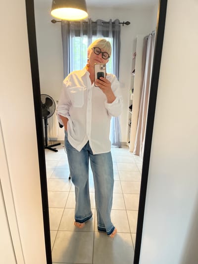 A woman with short blonde hair and eyeglasses is taking a selfie in a mirror. She is wearing a white button-down shirt and blue jeans, standing barefoot on a tiled floor. Behind her in the mirror's reflection is a room with a fan, a radiator, a window with grey curtains, and shelves with various items. The location is Chambœuf, France.