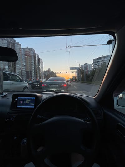 The image, taken from inside a car, shows a view of a city street at dusk. The sky is a soft orange and blue, suggesting sunset. The car appears to be in traffic, with other vehicles visible ahead. Buildings line the street, some appearing to be under construction with scaffolding. Telephone or power lines crisscross the sky. The interior of the car is visible, including the dashboard with a touchscreen display and the steering wheel. The display shows song information with the title "Don't Be Dumb" by ASAP Rocky and the time "21:24". License plates of some cars are visible, with one reading "Т225УВ24". The overall scene evokes a sense of urban life during a commute or travel.