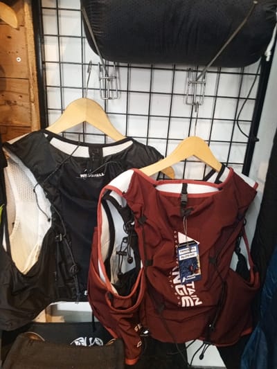 Two lightweight vests are displayed on a black wire grid wall, likely in a retail setting. On the left, a black vest with white mesh side panels and "WINDSHELL" branding is suspended from a light wooden hanger. On the right, a reddish-brown vest with a white collar trim is also on a light wooden hanger. This vest has a price tag attached, reading "Carex 10L", "Rp 250.000", and "MOUNTAIN RANGE ADVENTURE WITH". Both vests feature multiple pockets and elastic bungee cords for adjustment or storage. Above the vests, a dark, textured, cylindrical object is horizontally secured to the grid wall by metal clips. A vertical wooden panel is visible on the far left of the frame.