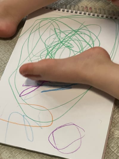 The image shows a child's foot resting on a white sheet of paper with colorful drawings on it. The drawings include a large green scribble, a few blue lines, and a purple circle. The child's foot is pale and has pink toes. The paper is part of a spiral bound notebook. The image is likely taken in a private residence in Moscow, Russia, based on the provided coordinates. The child's foot is resting on the paper, possibly while they are drawing. The action taking place is the child drawing, as evidenced by the colored lines on the paper. The child's foot is the main subject of the image. The environment appears to be indoors, likely a child's bedroom or playroom, as the image contains a carpeted floor and a notebook. The image is taken from a top-down perspective.
