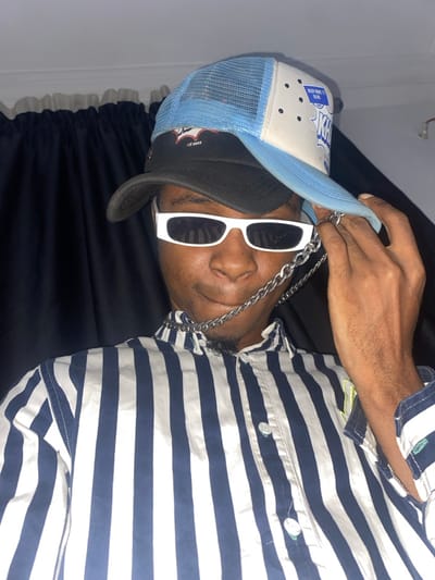 A man wearing a black and light blue baseball cap, white-framed sunglasses, and a silver chain is seen. He is wearing a blue and white striped short-sleeved shirt with visible buttons. He appears to be lying on a dark, possibly black, fabric, possibly a bed. The image is lit with moderate indoor lighting.