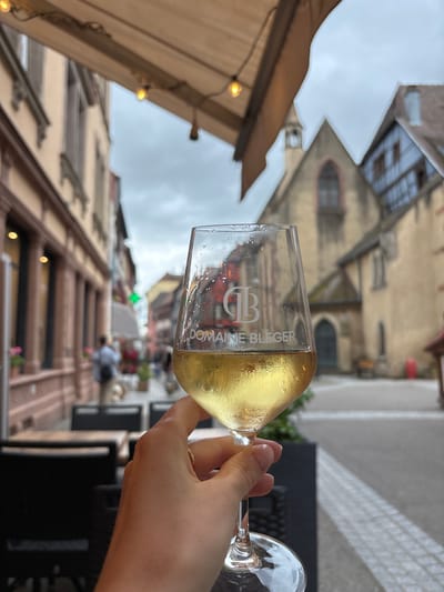 A close-up, low-angle shot shows a hand holding a partially filled wine glass, frosted with condensation. The glass displays a white logo with a stylized "B" and the text "DOMAINE BLEGER" below it, along with smaller text that is illegible. The background, blurred due to the shallow depth of field, depicts a street scene in Ribeauvillé, France, characterized by narrow buildings with traditional architecture, including timber-framed elements and a church spire. The sky is overcast, and an awning with string lights extends overhead, suggesting an outdoor seating area, possibly a restaurant or cafe. Several blurred figures are visible in the distance, walking along the street, which is paved with cobblestones. The overall impression is a casual moment of enjoyment in a picturesque European town.