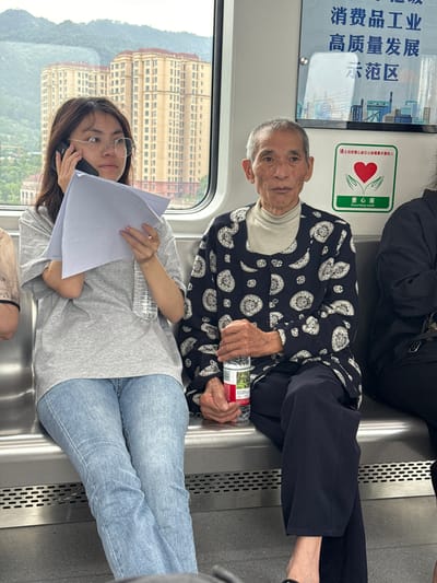 The image captures an indoor scene within a train, likely a subway car. An elderly Asian man in a patterned black and white jacket sits on the right, holding a water bottle. Next to him, a younger woman with glasses is on her phone while holding papers and another water bottle. Outside the window is a view of a city landscape featuring a multi-story tan building and green hills. Chinese text and symbols are on a sign in the train. The sign includes the text “Courtesy Seat” and Chinese text concerning high-quality industrial development and a demonstration zone. The people are fully dressed, and the scene appears to be ordinary public transportation. The location is Shuangfu, China.