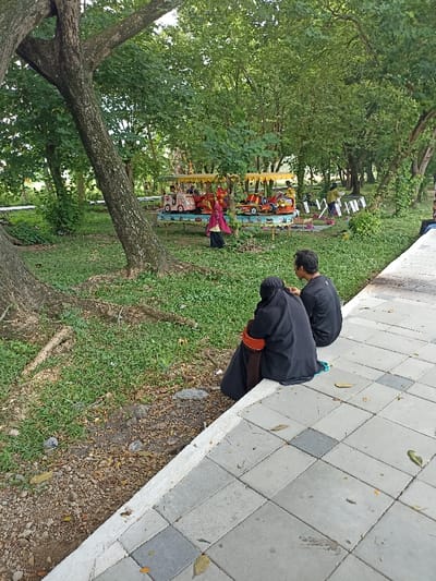 The image depicts a lively outdoor scene in a park or recreational area, characteristic of Mataram, Indonesia. In the foreground, two individuals are seated on a paved walkway comprised of grey and dark grey square tiles, facing away from the viewer. One person wears a black abaya or long dress with a matching head covering, featuring an orange accent on the arm. Beside them, another person is dressed in a black t-shirt.
Beyond the walkway, a grassy area with patches of exposed earth and fallen leaves leads to the main activity. A prominent tree trunk angles across the left side of the frame, indicating a well-established green space. In the middle ground, a colorful children's amusement ride is in operation. This ride consists of several car-shaped seats, some topped with yellow canopies, arranged in a circular or linear fashion, with multiple children visible enjoying the ride. A person in a red or pink garment stands near the ride, appearing to be an attendant or parent. Further in the background, a person in a light-colored top is seen amidst the dense green foliage of numerous trees, near some indistinct white structures.
The scene takes place during the daytime, with soft, diffused natural light filtering through the tree canopy, suggesting a pleasant and temperate climate suitable for outdoor leisure. The atmosphere is relaxed and family-oriented. No visible text can be discerned in the image.