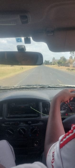 The scene is viewed from inside a vehicle traveling on a paved road in Njoro, Kenya. The driver's right arm, partially visible and wearing a white shirt with red shoulder stripes, grips the steering wheel. The vehicle's dashboard includes a radio unit, air vents, climate control knobs, and a green cord hanging. A rear-view mirror reflects a partial human profile. Outside the windshield, a two-lane asphalt road with yellow dashed center markings recedes into the distance under an overcast sky. The roadside features dry grassy verges, scattered trees, and distant structures on the right side of the road.