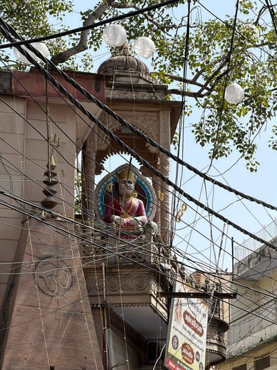 This is a low-angle, medium-shot photograph taken outdoors in the city of Delhi, India, during the daytime. The image is dominated by a complex network of tangled black electrical wires crisscrossing the frame. In the mid-ground, a statue of a deity, possibly Hindu, is visible within an ornate alcove on a reddish-brown stone structure. The deity is depicted wearing a golden crown and red garments, with arms resting in its lap. The structure itself features intricate carvings and is adorned with string lights. Above the alcove and draped over the branches of a tree, several white paper lanterns are suspended. The tree's leaves are green, and the sky is a clear blue. A poster, featuring images and text in Hindi, hangs from the structure on the right side of the frame. The overall impression is one of a densely urban environment with a strong religious and cultural presence.